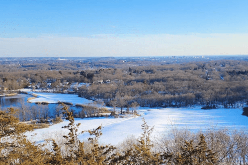 A scenic winter landscape featuring snow-covered fields, a lake, and distant city skyline under a clear blue sky.