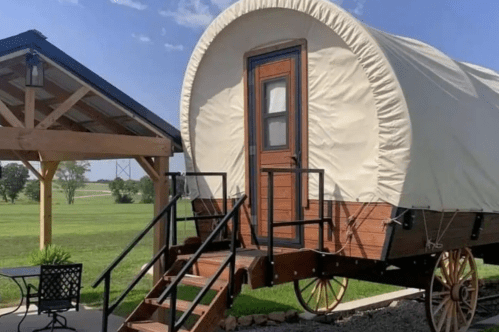 A covered wagon-style cabin with a wooden door, set in a grassy area near a pavilion.