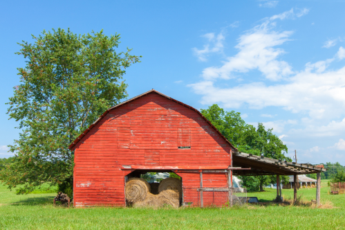 A red barn with hay bales under a roof, surrounded by green grass and trees under a blue sky.