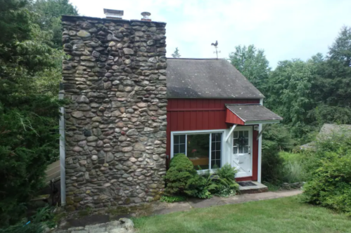 A small red and stone house surrounded by greenery, featuring a prominent stone chimney and a welcoming front entrance.