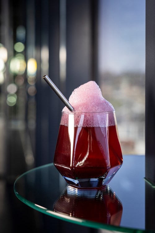 A vibrant pink drink with frothy foam, served in a geometric glass with a metal straw, on a reflective surface.