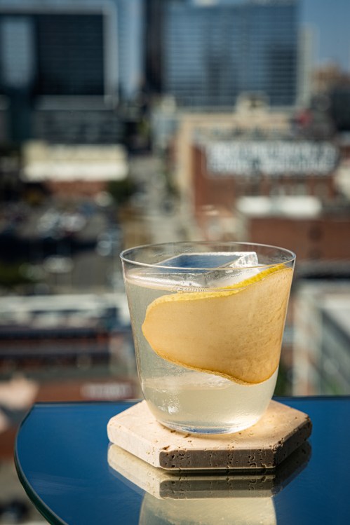 A refreshing cocktail with a pear slice sits on a coaster, overlooking a city skyline in the background.