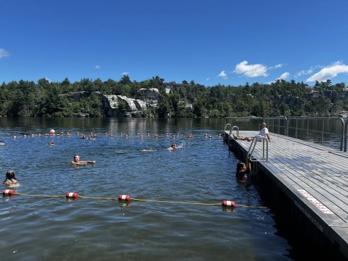 A sunny day at a lake with swimmers and a wooden dock, surrounded by trees and rocky cliffs in the background.