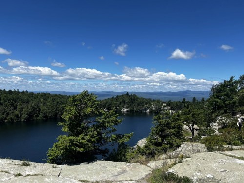 A scenic view of a lake surrounded by trees and mountains under a blue sky with fluffy clouds.