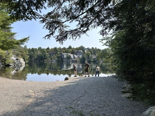 Three people stand by a calm lake surrounded by trees and rocky cliffs on a sunny day.