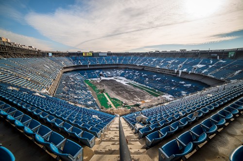 An empty stadium with blue seats and a partially covered field, under a cloudy sky.