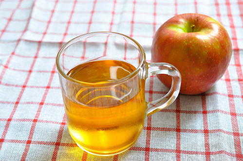 A clear glass cup of golden liquid next to a red apple, set on a checkered tablecloth.