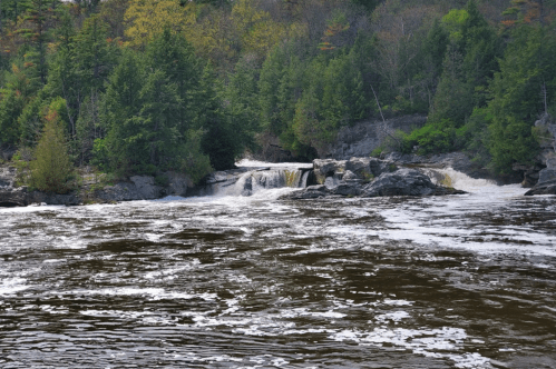 A serene waterfall cascades over rocks, surrounded by lush green trees and a flowing river under a clear sky.