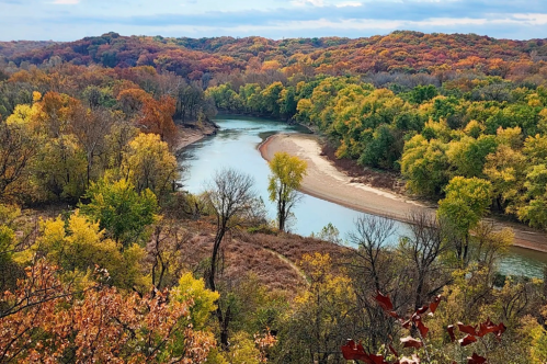 A winding river flows through a vibrant landscape of autumn foliage in various shades of orange, yellow, and green.