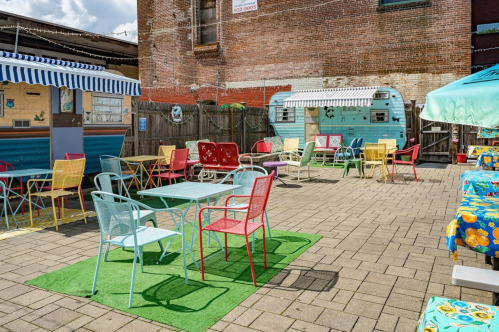 Colorful outdoor seating area with tables and chairs, surrounded by a brick wall and vintage trailers. Bright and inviting atmosphere.