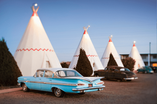 Vintage cars parked near iconic teepee-shaped motels at dusk, with soft lighting illuminating the scene.