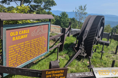 Sign about the Old Cable Railway with a large wheel, set against a scenic mountain backdrop.