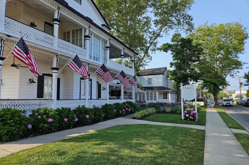A charming white house with American flags, surrounded by greenery and flowers, on a sunny street.