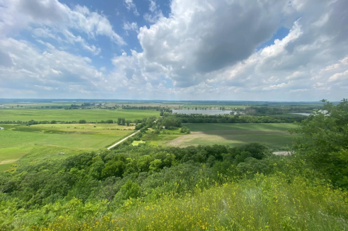 A panoramic view of lush green fields and a river under a partly cloudy sky.