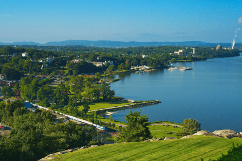 A scenic view of a river with lush greenery, a train along the shore, and distant mountains under a clear blue sky.