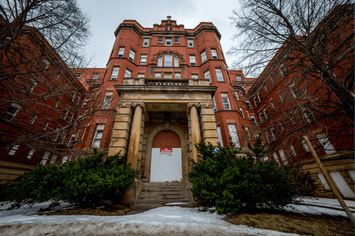 Historic red brick building with a grand entrance, surrounded by bare trees and patches of snow.
