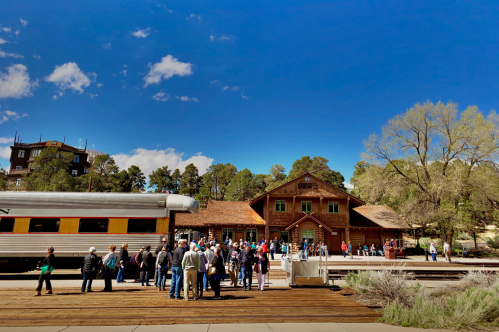 A crowd gathers near a vintage train station with a wooden building and clear blue sky in the background.