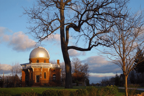 A historic observatory with a silver dome, surrounded by trees and a grassy area under a cloudy sky.