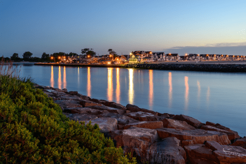 A serene waterfront scene at dusk, with reflections of lights on calm water and rocky shoreline.