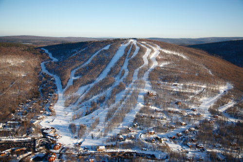 Aerial view of a snow-covered ski slope with winding trails and nearby houses nestled in a wooded area.