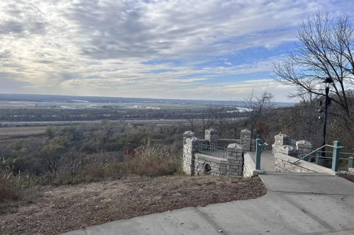 Scenic view from a hilltop overlooking a river valley, with a pathway and stone railing in the foreground.
