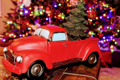 A vintage red toy truck with a Christmas tree in the back, set against a colorful, decorated Christmas tree backdrop.