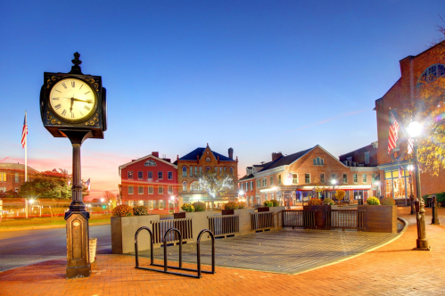 A historic town square at dusk, featuring a clock, brick pathways, and charming buildings with festive decorations.