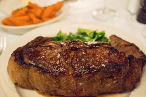 A perfectly cooked steak with a crispy crust, served with sautéed greens and a side of carrots on a white plate.