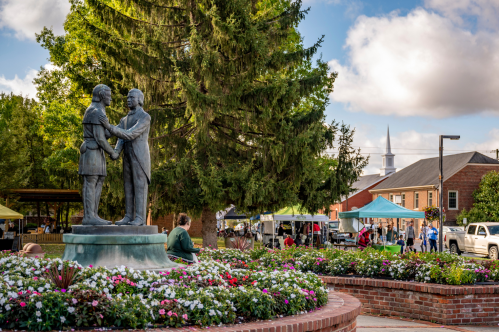 A statue of two men shaking hands surrounded by flowers, with a market and buildings in the background.