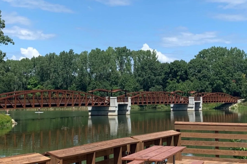 A scenic view of a metal bridge spanning a calm river, surrounded by lush green trees and a clear blue sky.