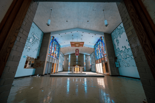 Interior of an abandoned church with stained glass windows, peeling walls, and a central altar. Light filters through the glass.