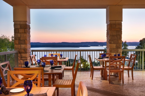 A scenic outdoor dining area with wooden tables and blue glassware, overlooking a sunset by the water.