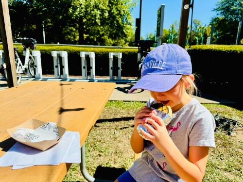 A young girl in a blue cap enjoys a snack at a picnic table on a sunny day, with bikes in the background.
