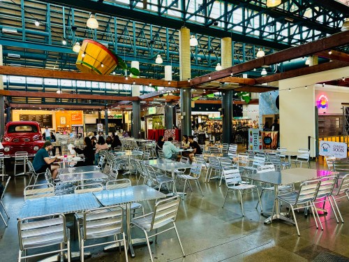A spacious indoor market with metal tables, people dining, and colorful stalls in the background under a high ceiling.