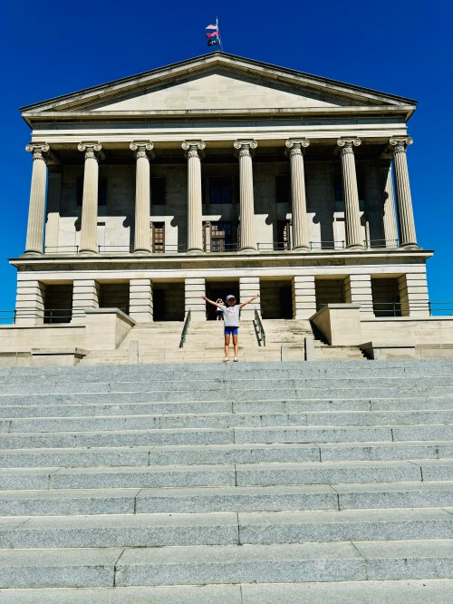 A person stands with arms outstretched at the base of a grand stone building with columns and a flag on top.