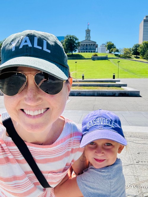 A woman and a child smile for a selfie, wearing hats, with a grassy area and a building in the background.