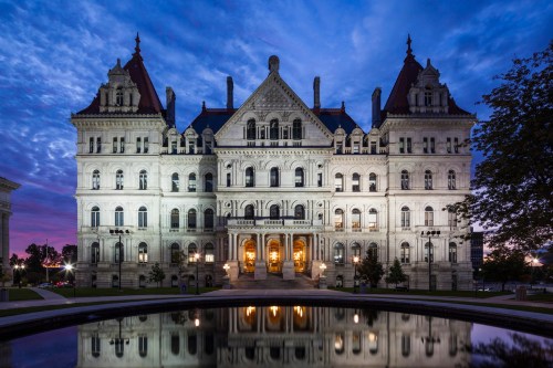 The New York State Capitol building illuminated at dusk, reflecting in a nearby pond under a colorful sky.