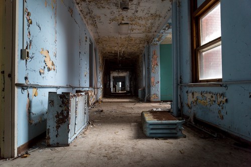 An abandoned hallway with peeling blue paint, debris on the floor, and old radiators along the walls.