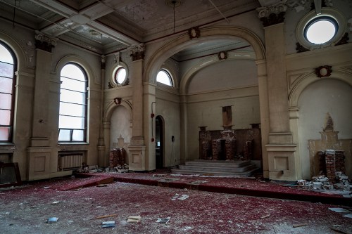 An abandoned room with peeling walls, arched windows, and a dusty red carpet, featuring a crumbling altar at the back.