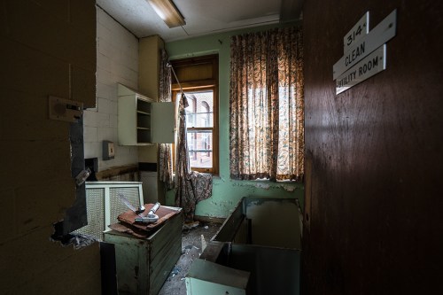 Abandoned utility room with peeling paint, cluttered surfaces, and sunlight filtering through dusty curtains.