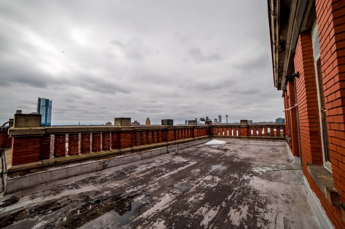 A rooftop view featuring a cloudy sky, brick walls, and a worn surface, with distant city buildings visible.