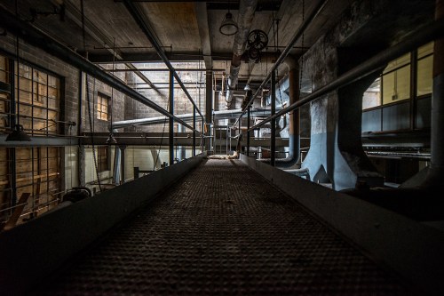 A dimly lit industrial space with metal walkways, pipes, and large windows, creating a gritty, atmospheric scene.