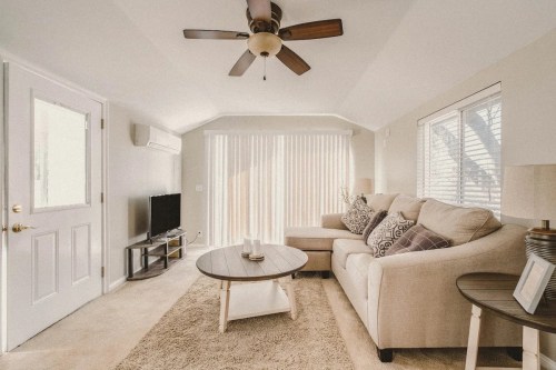 Cozy living room with a beige sofa, coffee table, TV, and large windows with vertical blinds. Light and airy atmosphere.