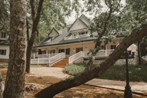 A large, elegant house with a wraparound porch, surrounded by trees and greenery.
