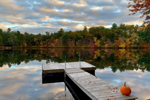 A serene lake scene with a wooden dock, colorful autumn trees, and a small pumpkin on the shore under a cloudy sky.