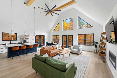 Modern living room with large windows showcasing autumn trees, featuring a brown leather sofa, green couch, and a kitchen bar.