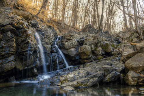A serene waterfall cascades over rocky terrain, surrounded by trees in a tranquil forest setting.