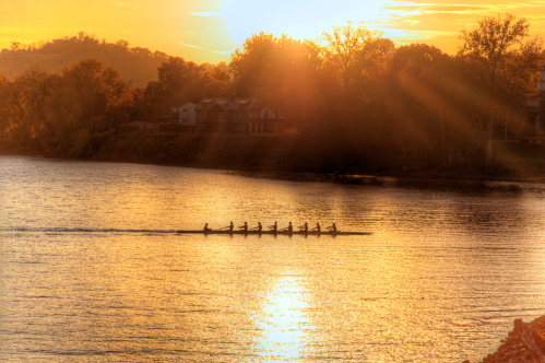 A rowing team glides across a shimmering river at sunset, surrounded by trees and a warm golden glow.