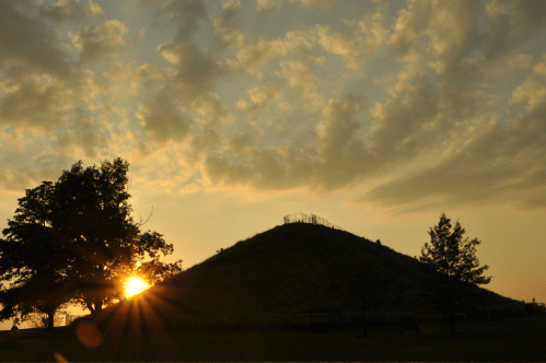 Silhouette of a hill at sunset, with trees in the foreground and a cloudy sky.