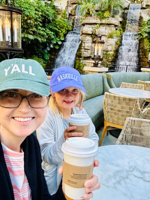 A woman and a girl hold coffee cups, smiling in front of a lush backdrop with waterfalls and outdoor seating.